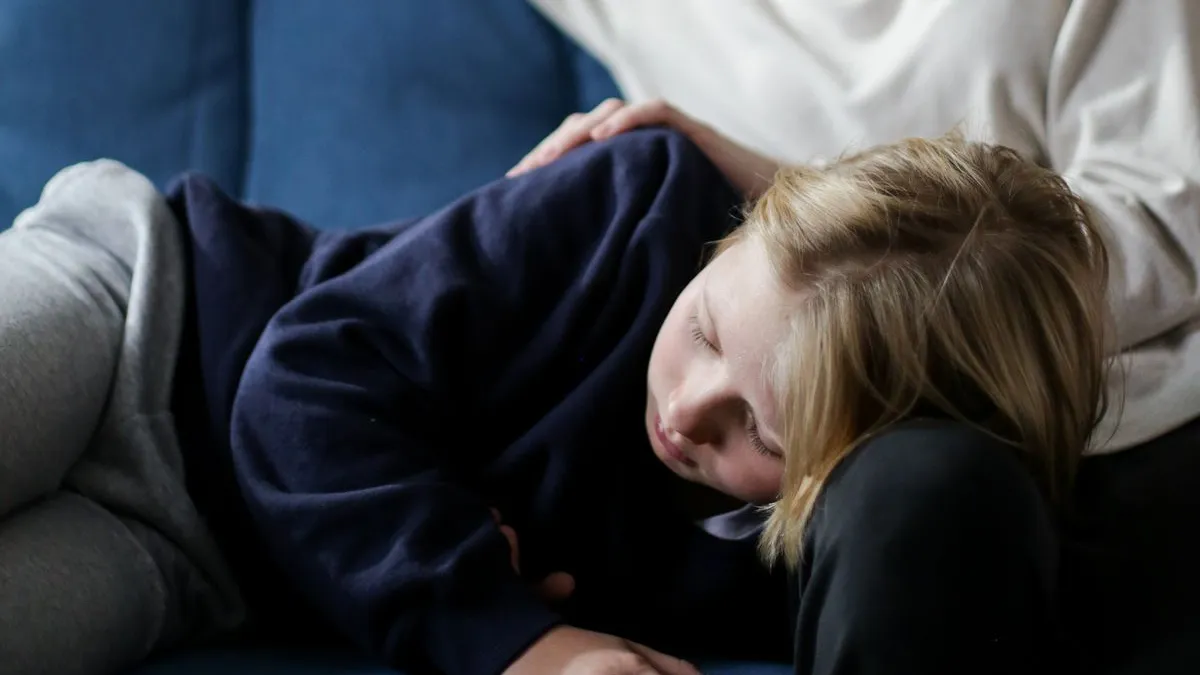 Kind liegt zusammengerollt auf dem Sofa, ein Elternteil hält die Hand auf seiner Schulter