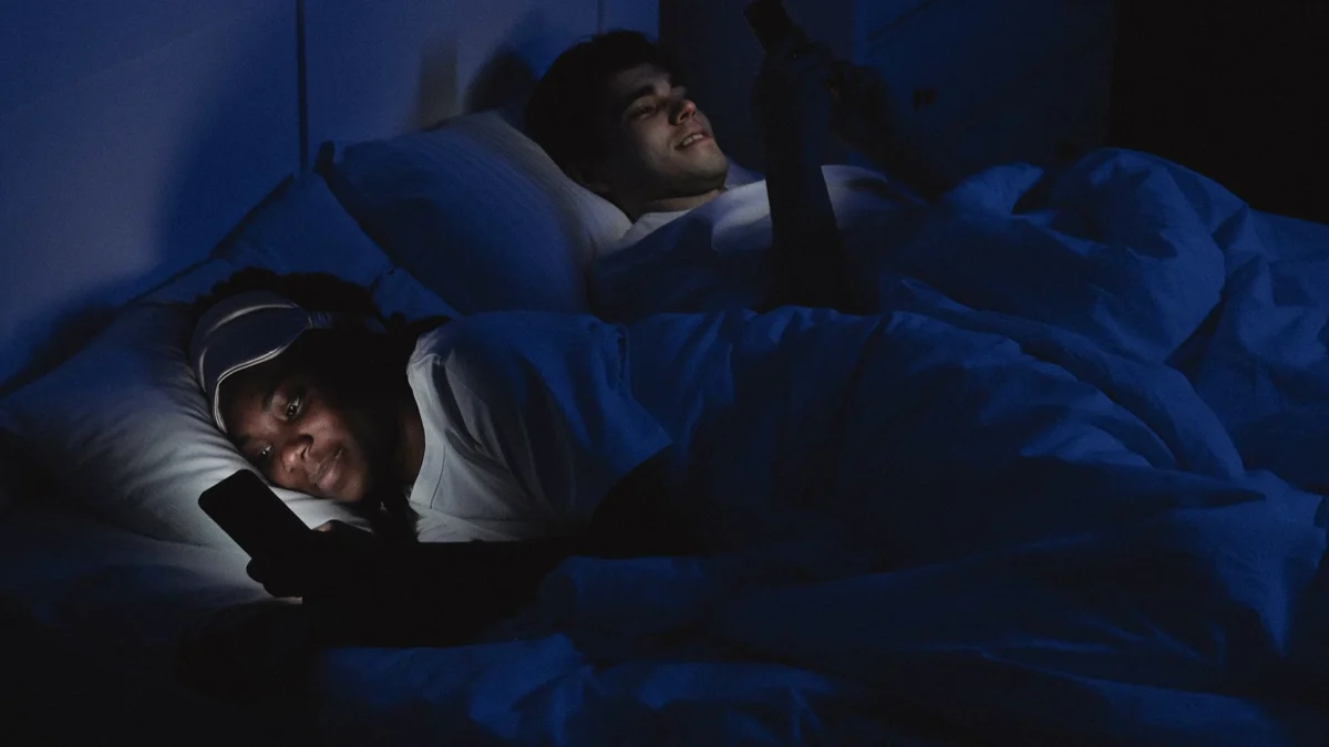 Paar liegt nachts im Bett, beide voneinander abgewandt am leuchtenden Smartphone