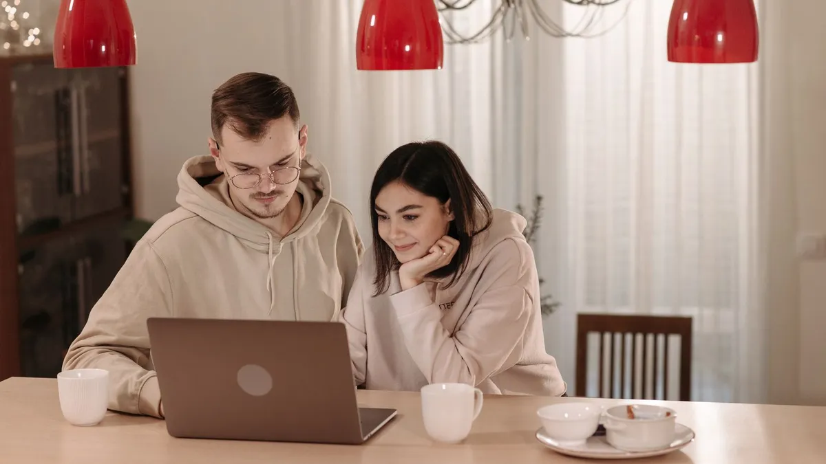 Paar in Kapuzenpullovern sitzt am Küchentisch und schaut gemeinsam auf einen Laptop