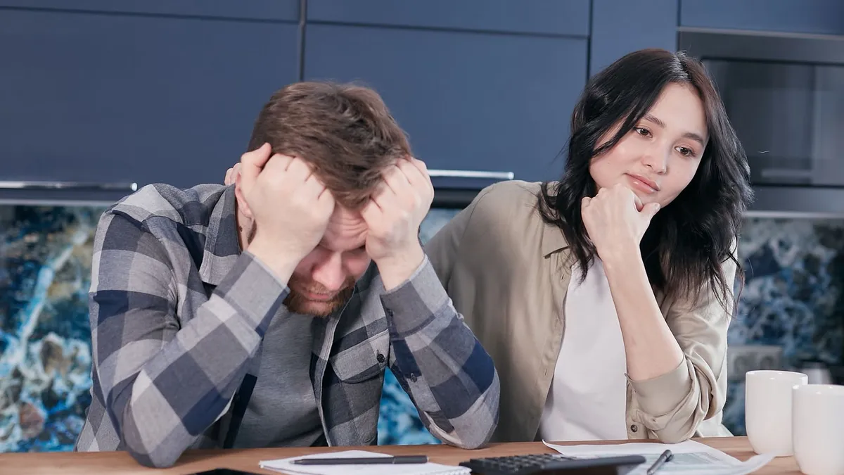 Mann vergräbt den Kopf in den Händen, seine Partnerin sitzt besorgt neben ihm am Küchentisch
