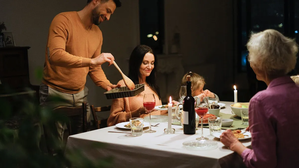 Familie beim Abendessen bei Kerzenlicht, ein Mann schöpft Essen auf, am Tisch sitzen eine Frau, ein Kind und die Großmutter