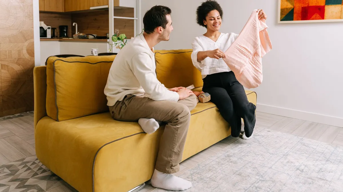 Frau hält auf einem gelben Sofa ein rosafarbenes Kleidungsstück hoch, der Mann schaut zu