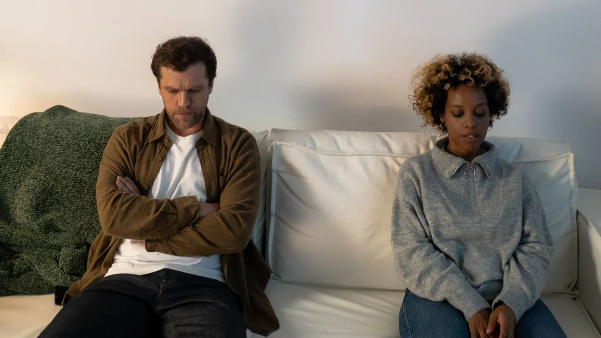 Mann mit verschränkten Armen und Frau mit gesenktem Blick sitzen mit Abstand nebeneinander auf einem weißen Sofa