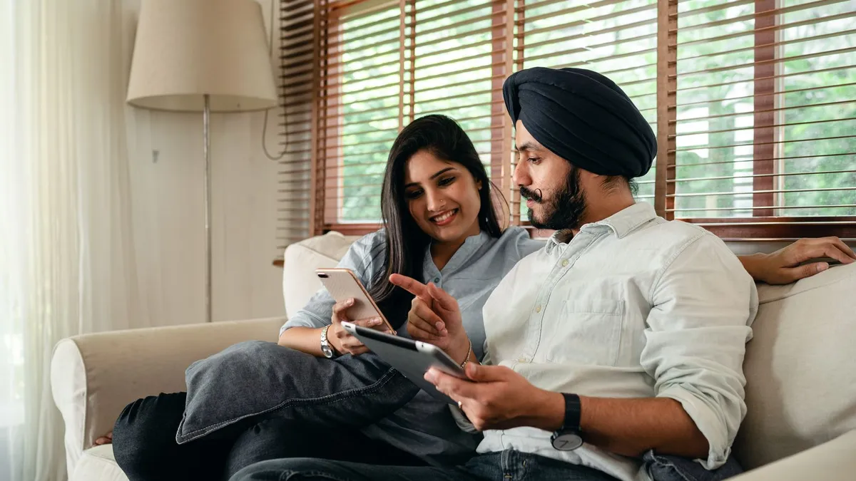 Frau mit Smartphone und Mann mit Tablet sitzen lächelnd nebeneinander auf einem Sofa