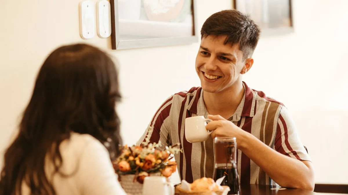 Mann im gestreiften Hemd lächelt eine Frau mit dunklen Haaren an einem Café-Tisch an