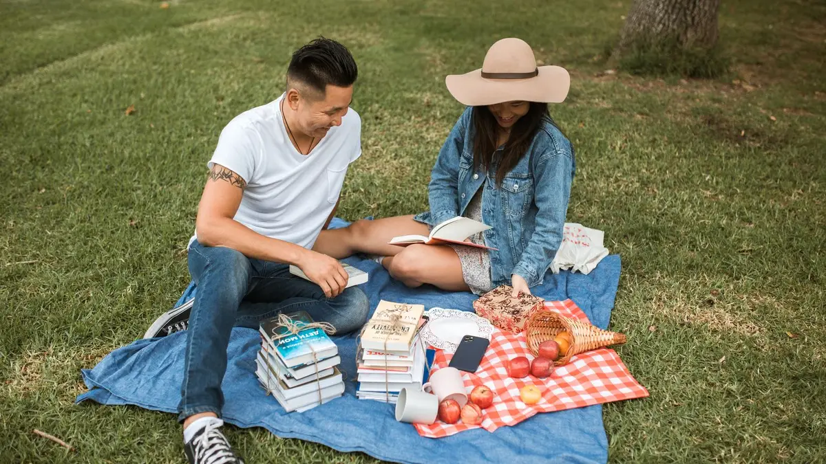Mann und Frau lesen auf einer Picknickdecke im Gras, neben ihnen liegen Bücherstapel und Äpfel