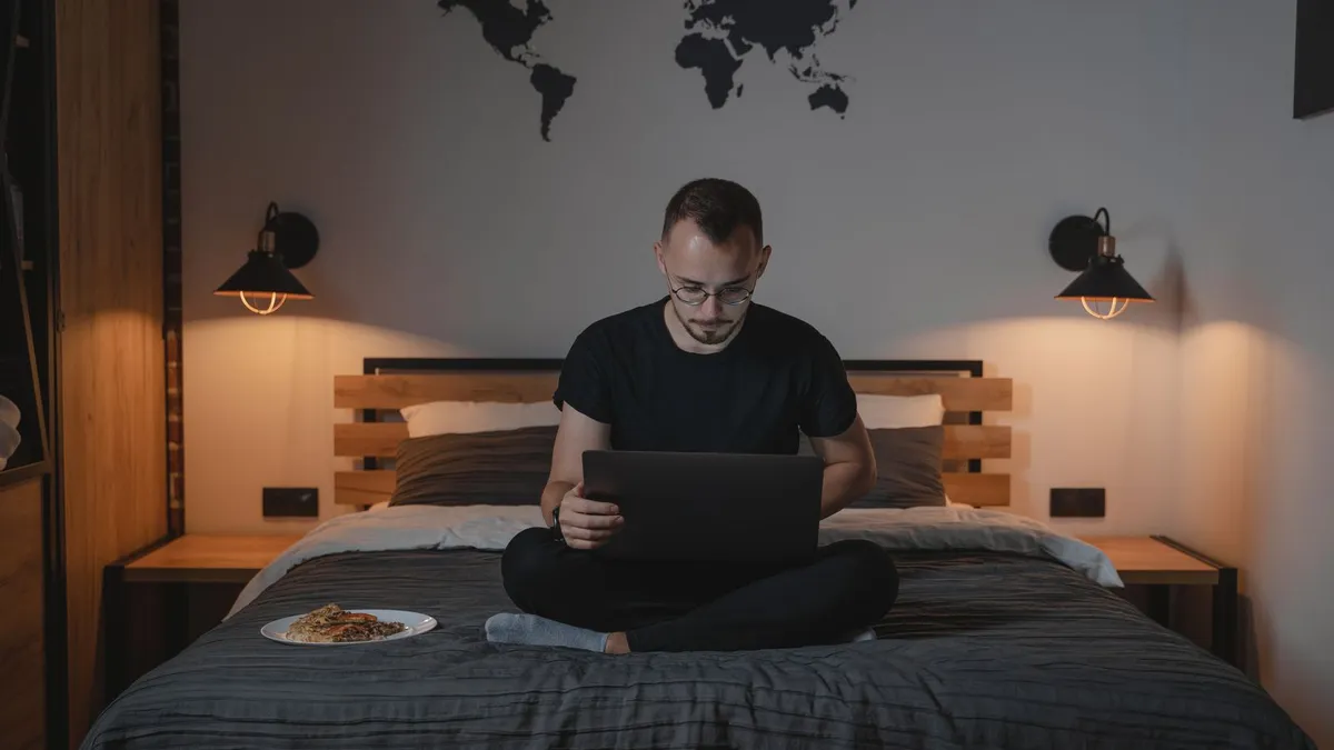 Ein Mann sitzt abends im Schneidersitz auf dem Bett und schreibt auf seinem Laptop