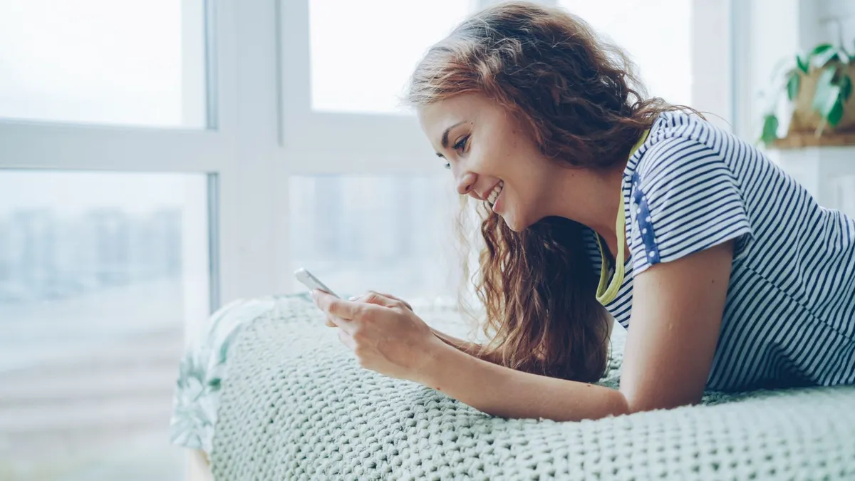 Eine junge Frau liegt am Fenster auf dem Bett und schreibt lächelnd auf ihrem Smartphone