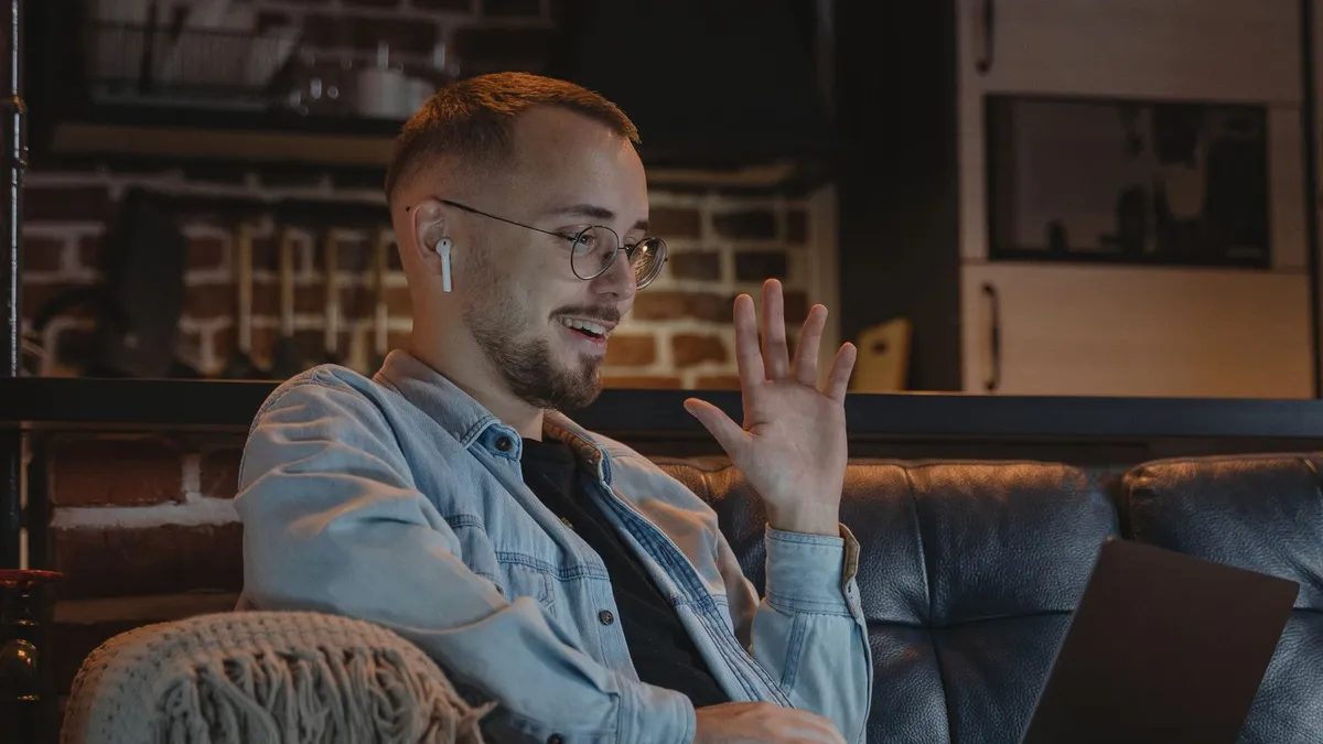 Ein Mann mit Brille und Kopfhörer im Ohr winkt lächelnd in die Kamera seines Laptops