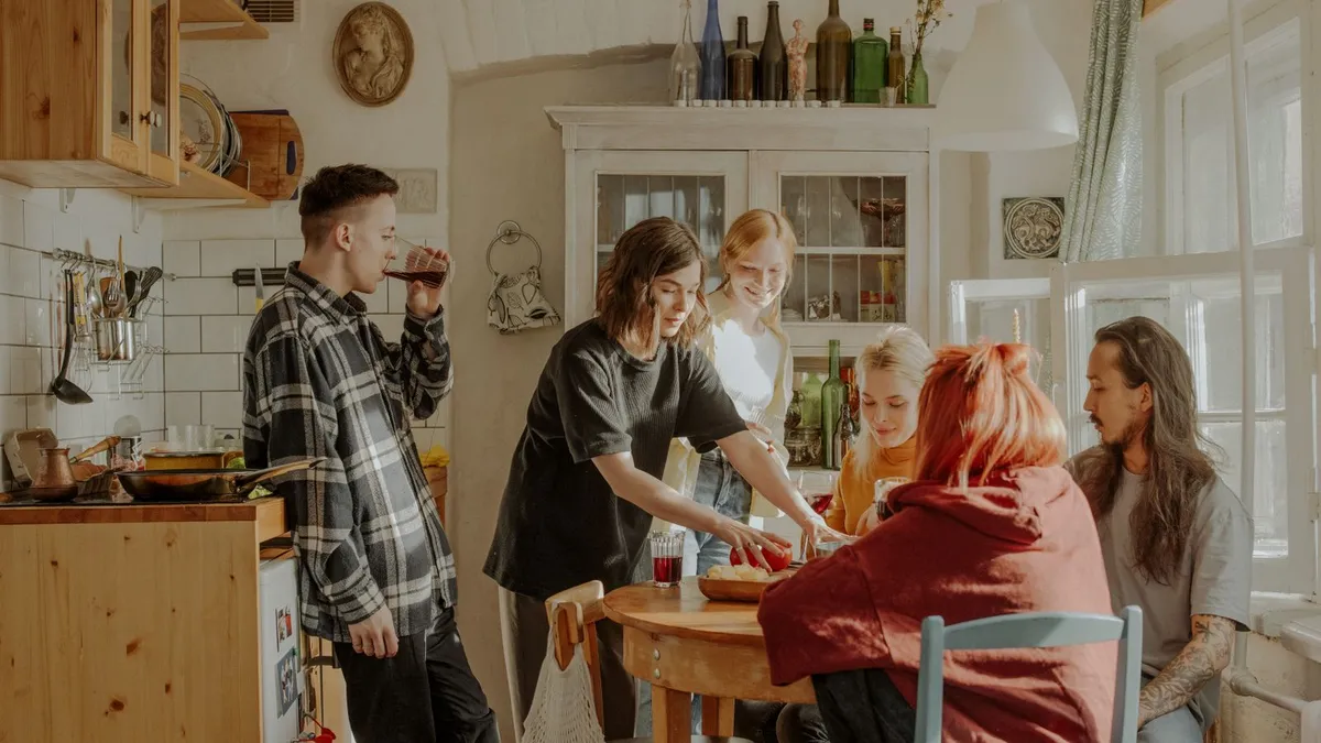 Mehrere junge Menschen stehen und sitzen in einer hellen Gemeinschaftsküche mit Holzmöbeln
