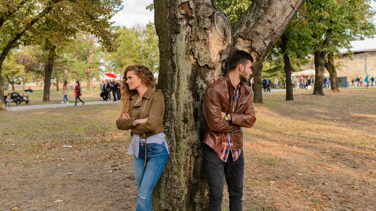 Frau und Mann lehnen mit verschränkten Armen an gegenüberliegenden Seiten eines Baumstamms in einem Herbstpark
