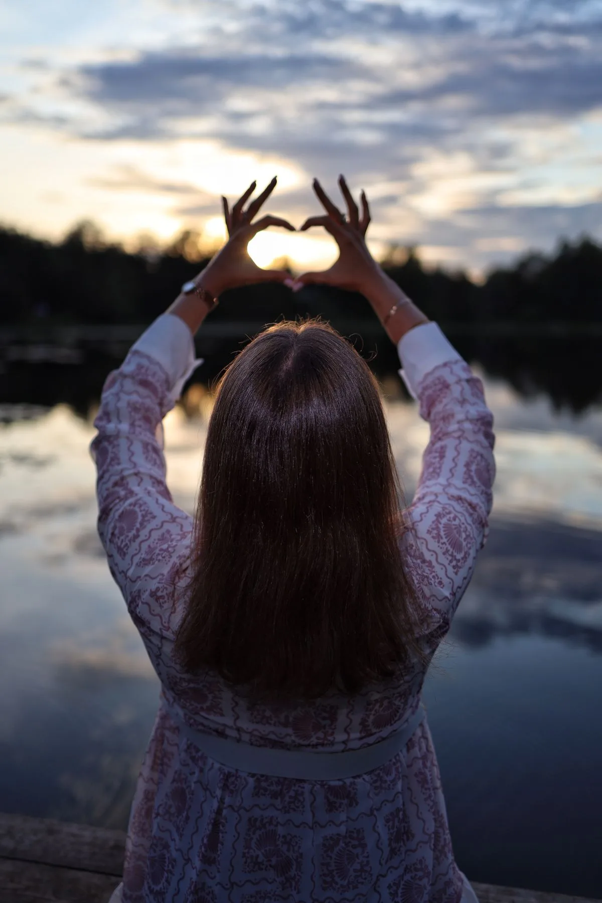Frau formt mit ihren Händen ein Herz vor dem Sonnenuntergang über einem ruhigen See