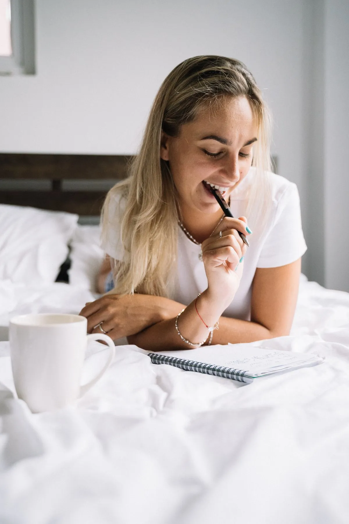 Frau liegt lächelnd mit Notizbuch und Stift auf dem Bett, daneben eine Tasse Kaffee