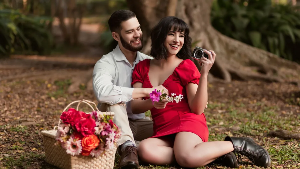 Paar sitzt bei einem Picknick im Park, sie hält eine Kamera, daneben steht ein Korb mit Blumen