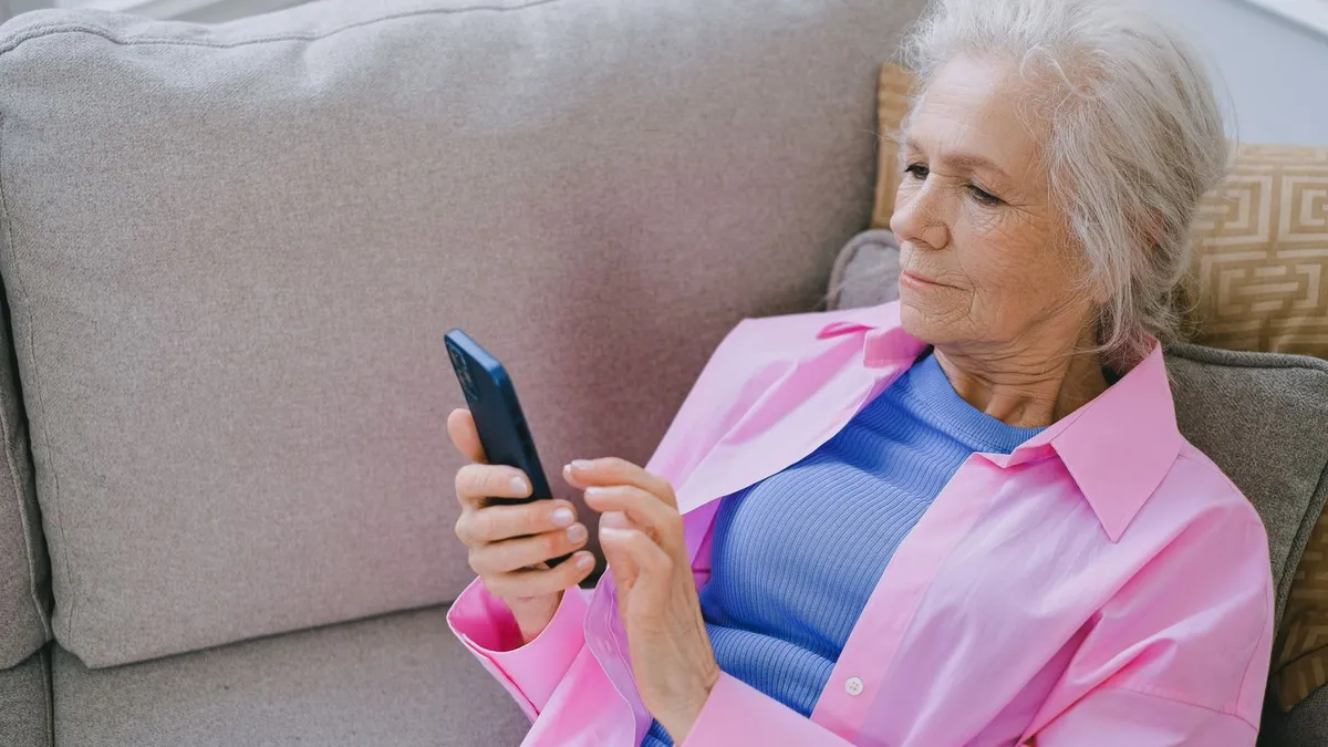Frau mit grauen Haaren und rosa Bluse liegt auf dem Sofa und tippt auf ihrem Smartphone
