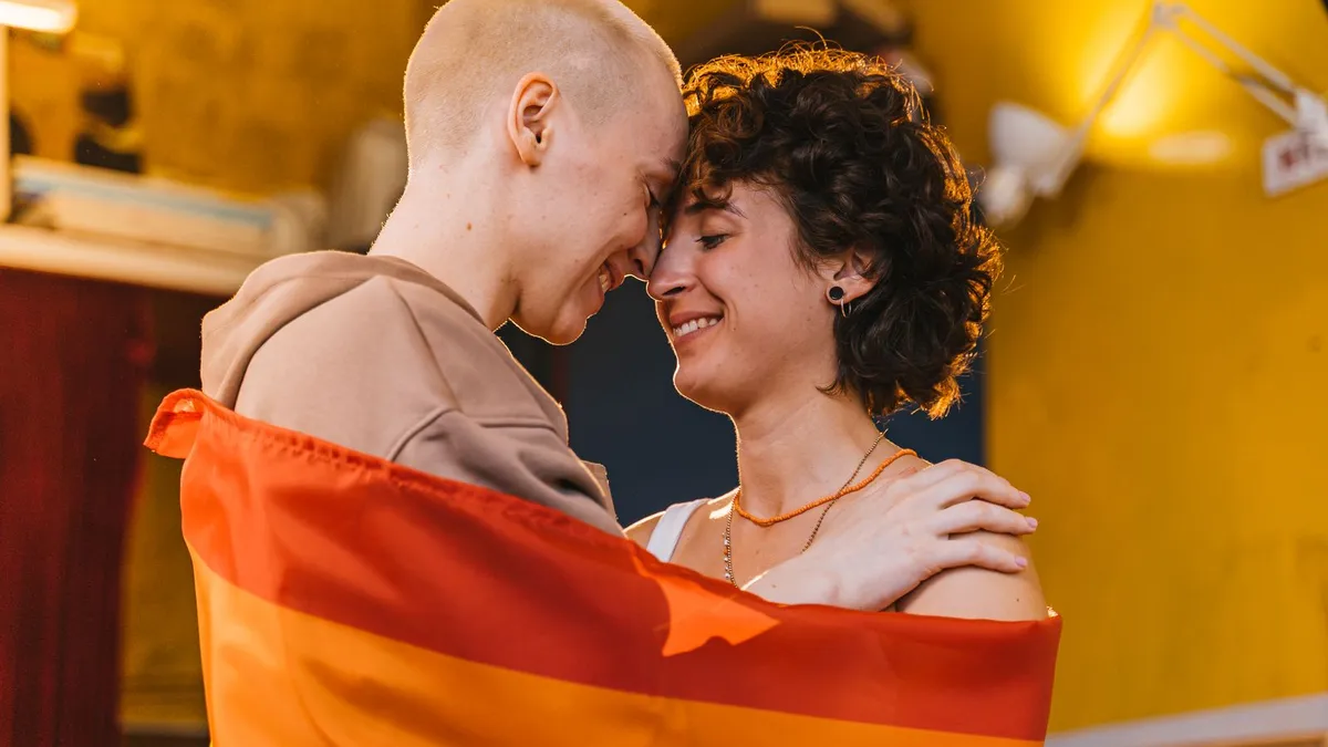 Zwei Menschen berühren sich lächelnd mit der Stirn, um beide ist eine Regenbogenflagge gelegt