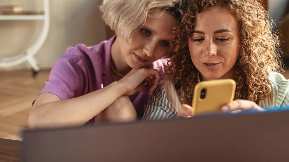 Zwei Frauen schauen hinter einem Laptop gemeinsam auf ein gelbes Smartphone