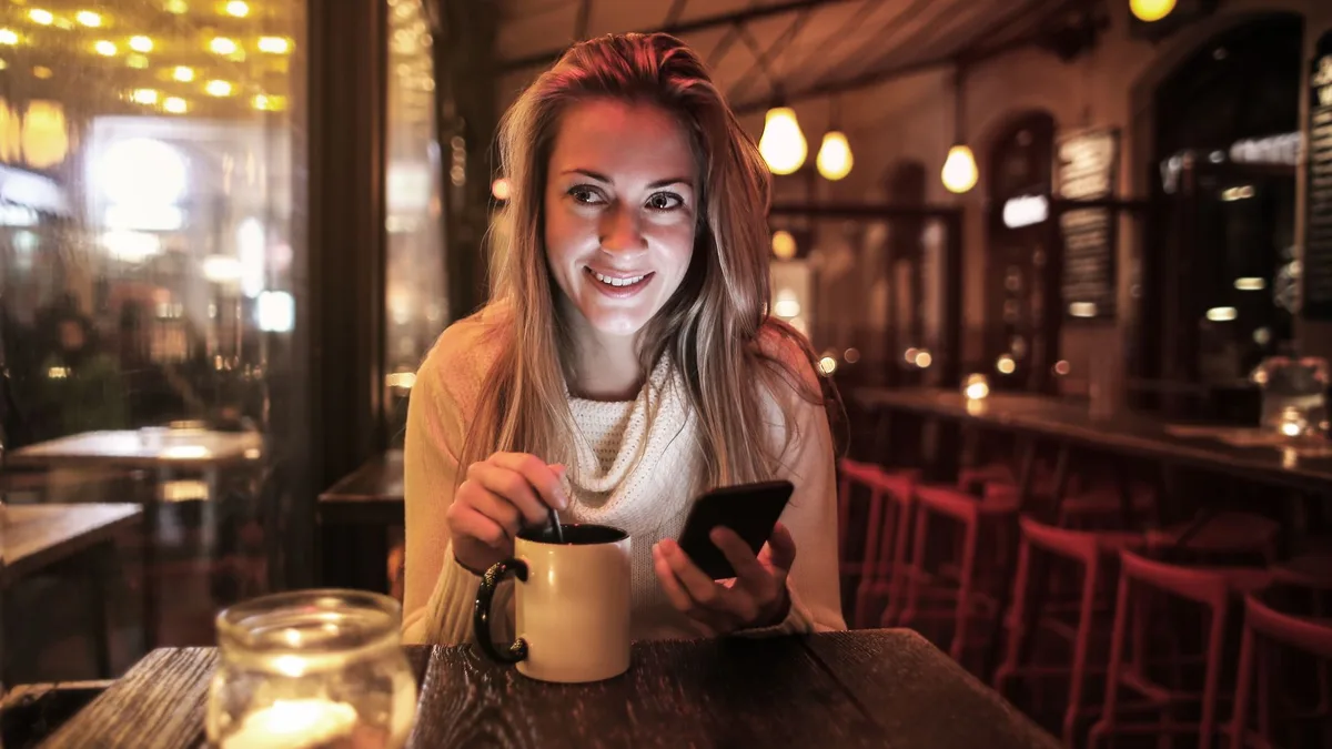 Lächelnde Frau mit Kaffeetasse und Smartphone am Abend in einem Café