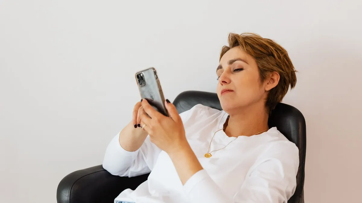 Frau mit kurzem Haar sitzt in einem Sessel und scrollt mit müdem Blick auf ihrem Smartphone