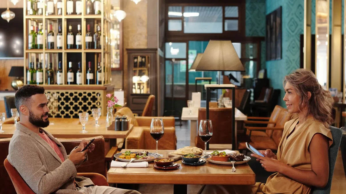 In einem Restaurant sitzen ein Mann und eine Frau am gedeckten Tisch, beide halten ein Smartphone in der Hand