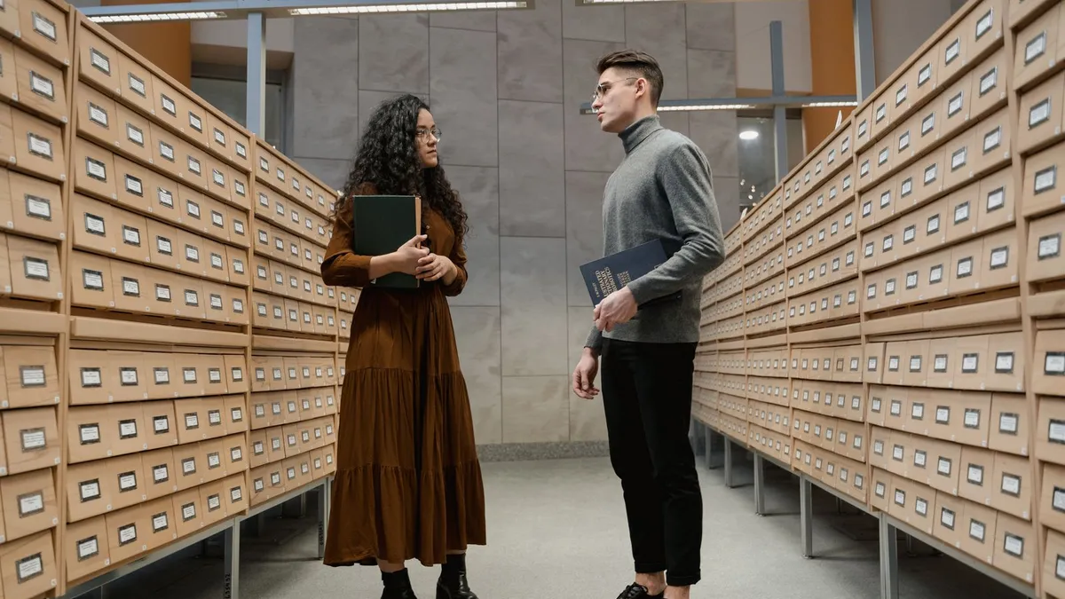 Frau und Mann mit Büchern in der Hand stehen sich in einem Gang mit Karteikästen einer Bibliothek gegenüber
