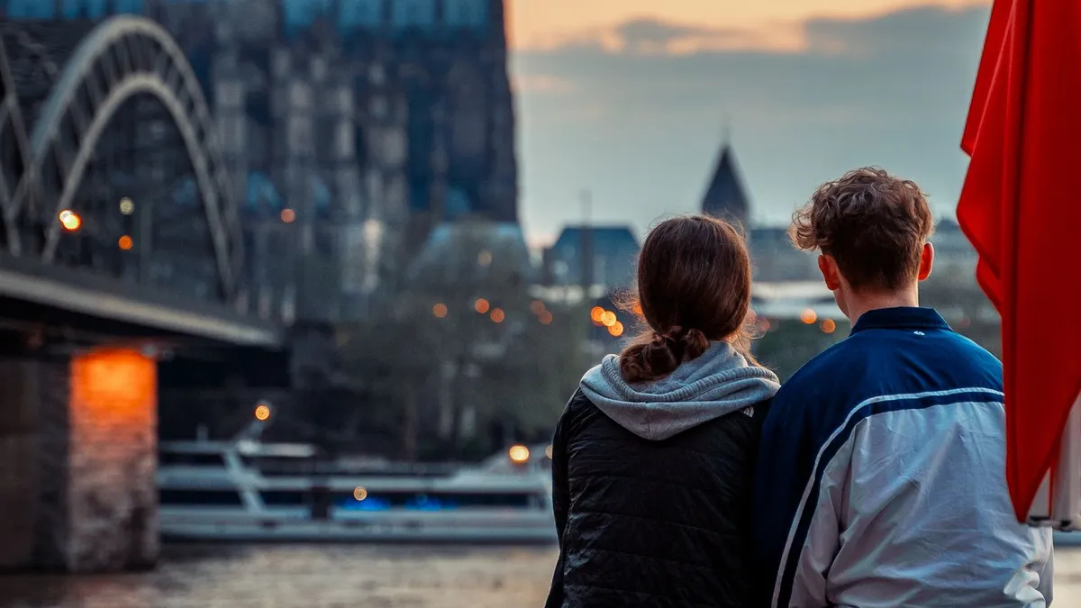 Junges Paar sitzt am Rheinufer in Köln und blickt in der Abenddämmerung auf die Hohenzollernbrücke und den Dom