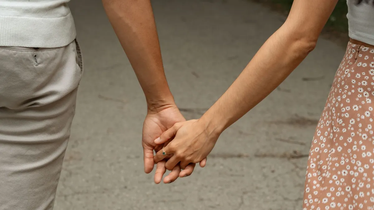 Zwei Menschen gehen Hand in Hand einen Weg entlang, zu sehen sind nur Arme, Hände und Kleidung