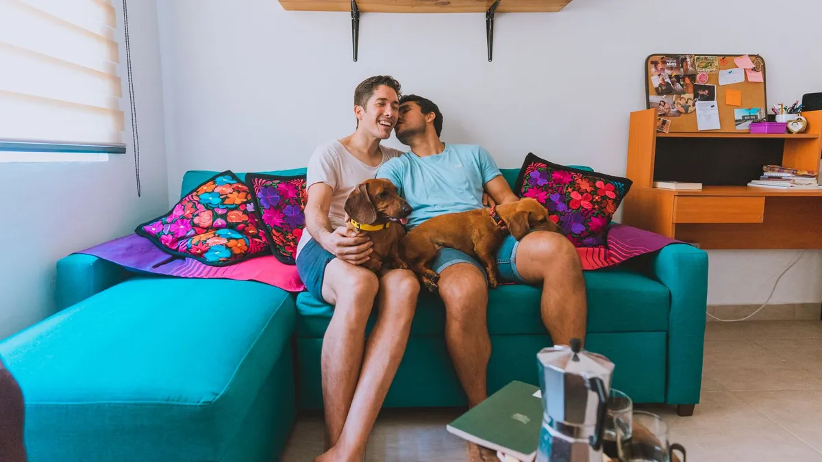 Zwei Männer sitzen lachend und eng beieinander auf einem Sofa, bei ihnen zwei Dackel