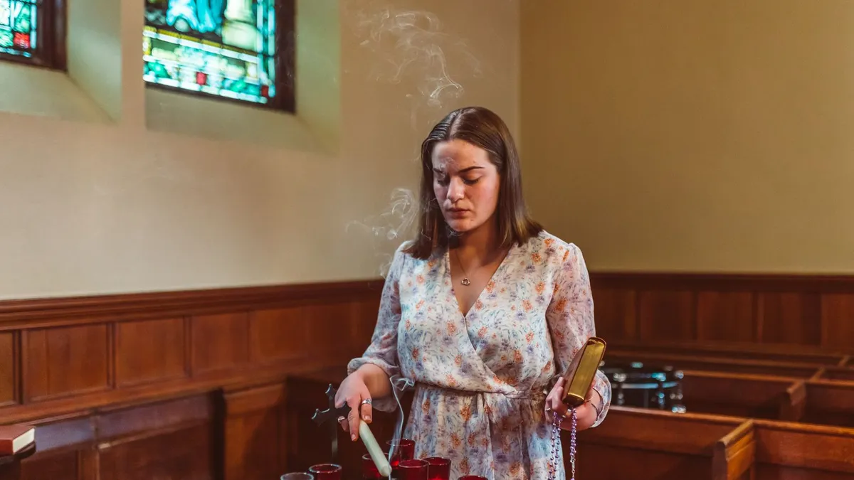 Frau zündet in einer Kirche eine Kerze an, im Hintergrund Buntglasfenster und Kirchenbänke