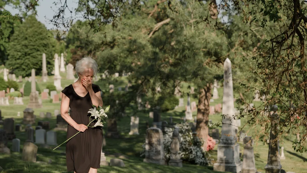 Ältere Frau in dunklem Kleid steht mit weißen Lilien zwischen Grabsteinen auf einem Friedhof