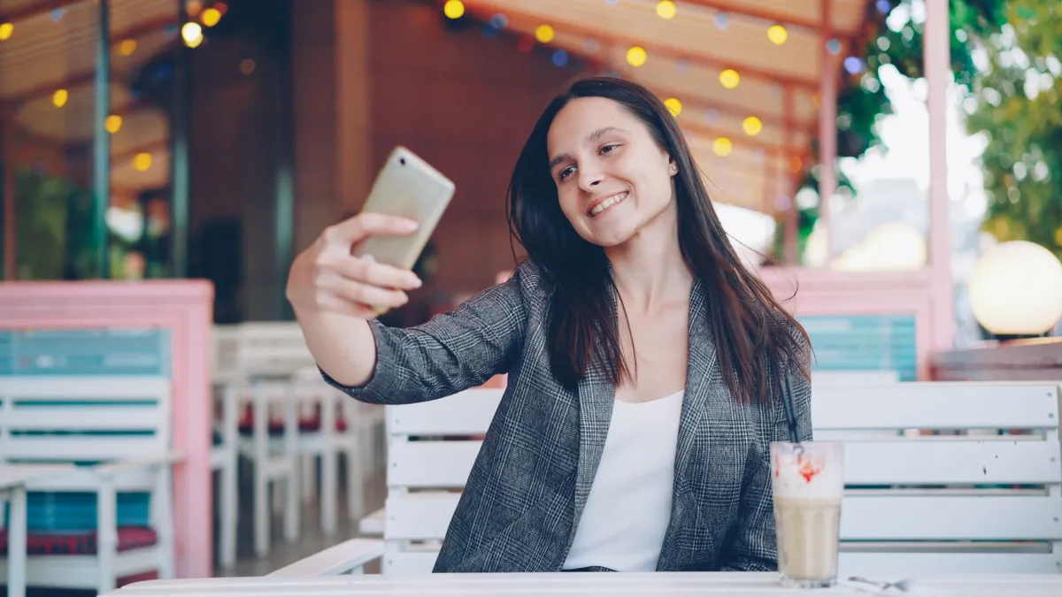 Lächelnde Frau mit langen dunklen Haaren macht in einem Café ein Selfie mit ihrem Smartphone