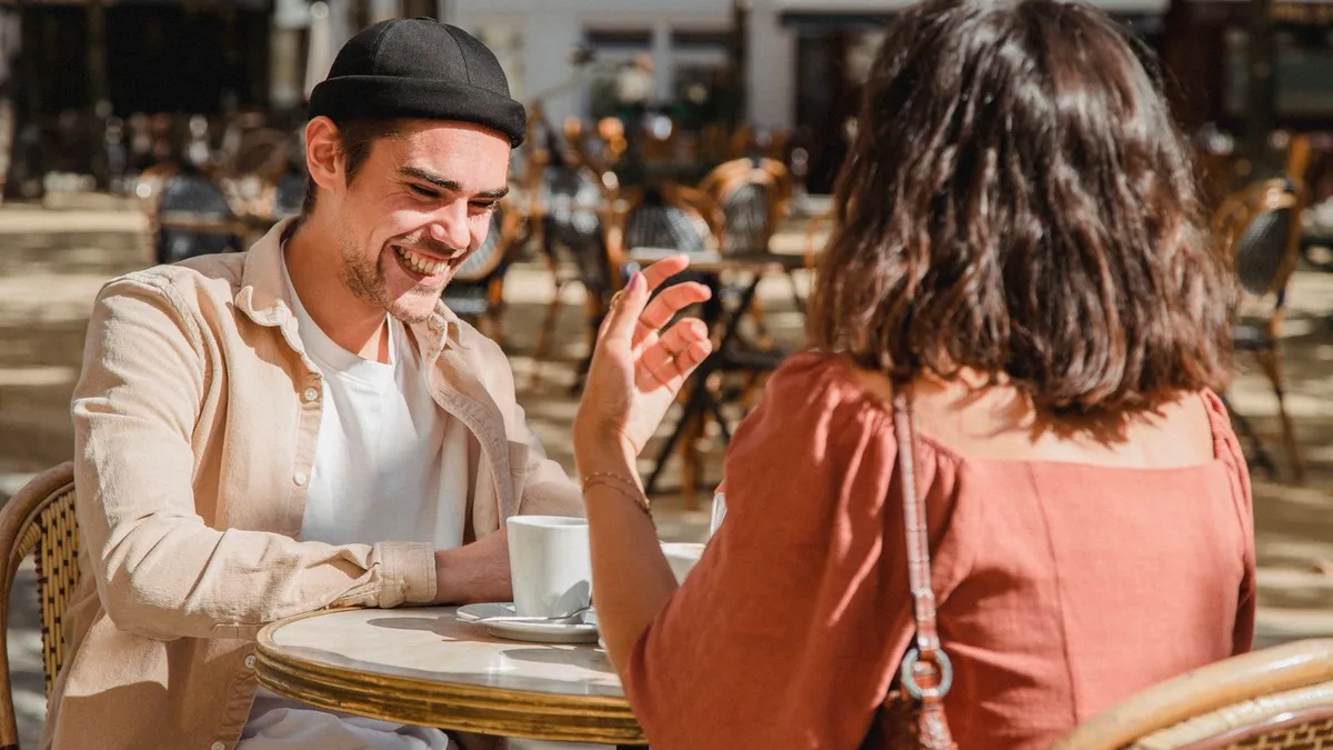 Lachender Mann mit Mütze gestikuliert an einem Café-Tisch im Freien, ihm gegenüber eine Frau von hinten
