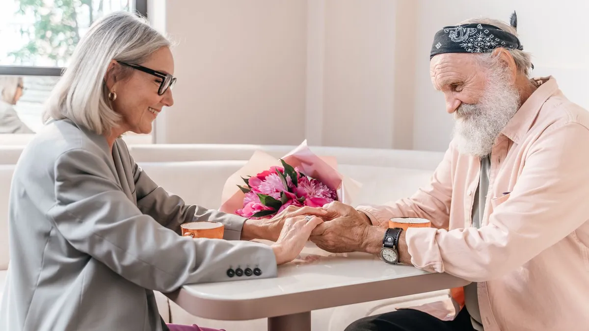 Älteres Paar hält sich an den Händen an einem Tisch, zwischen ihnen ein Strauß pinker Blumen