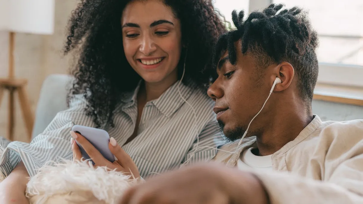 Frau und Mann sitzen auf einem Sofa und hören mit Kopfhörern gemeinsam Musik vom Smartphone