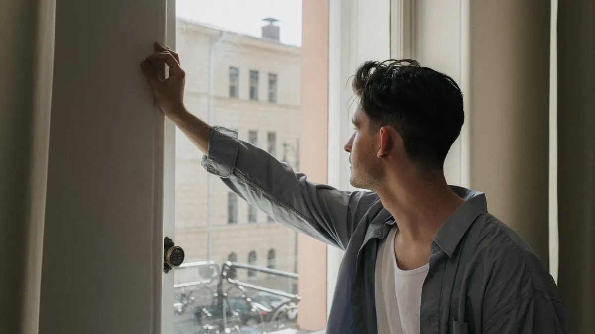 Mann lehnt am Fensterrahmen und blickt abwesend nach draußen auf eine Häuserfassade