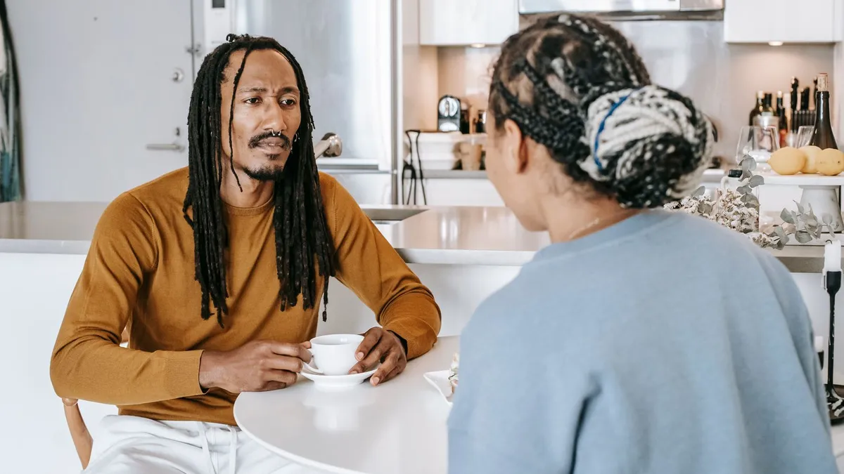 Mann mit Dreadlocks hält eine Tasse und spricht ernst mit einer Frau, die ihm am Küchentisch gegenübersitzt