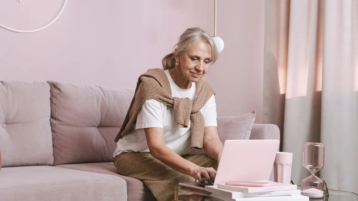 Frau mit grauem Haar sitzt lächelnd auf dem Sofa und tippt auf einem Laptop