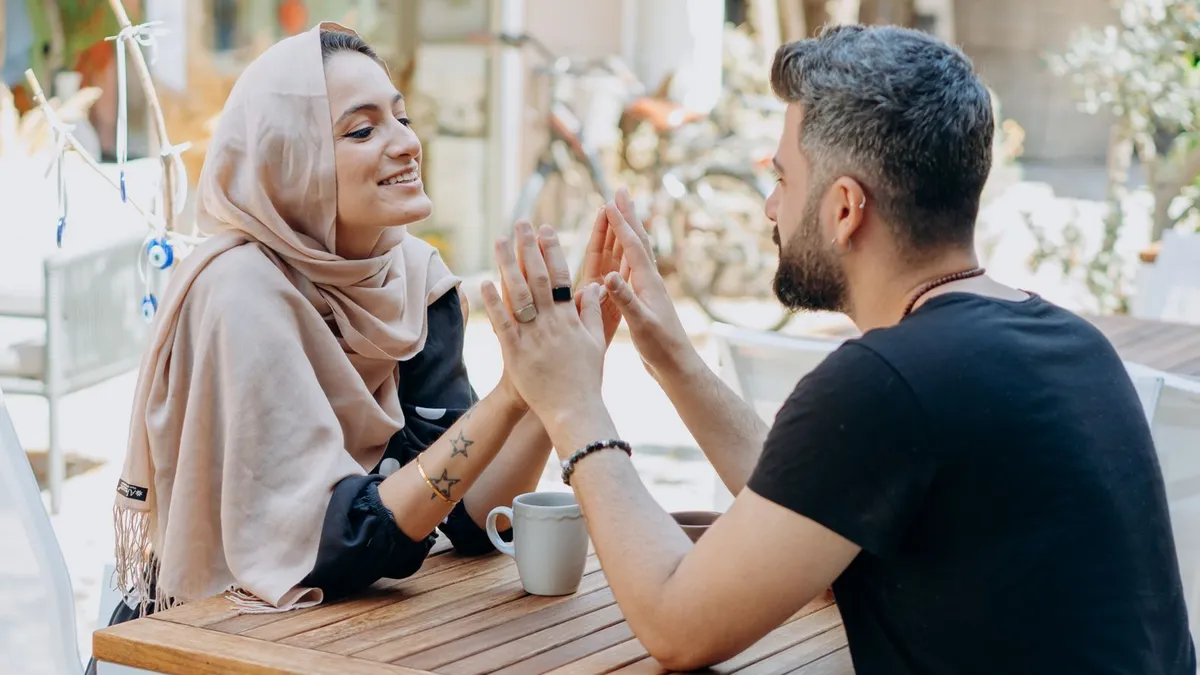 Frau mit Kopftuch und Mann legen an einem Café-Tisch die Handflächen aneinander