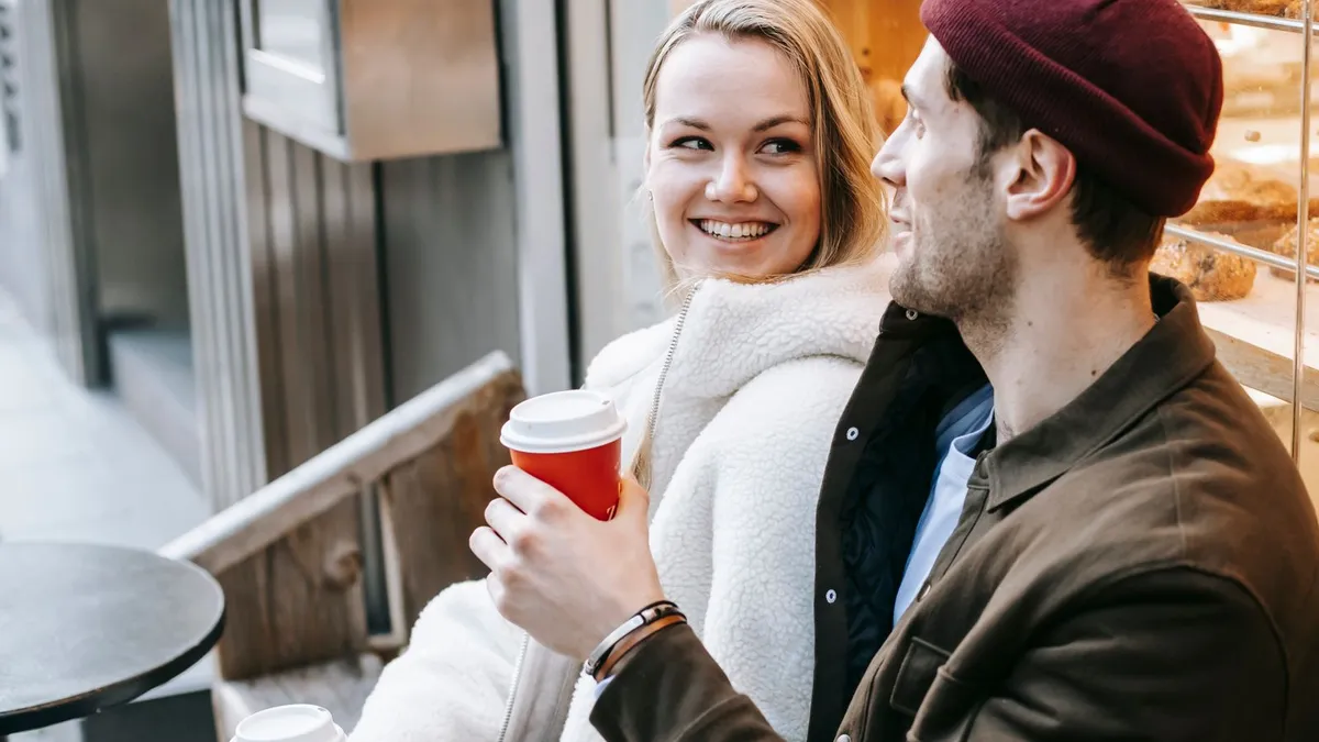 Lächelnde Frau mit rotem Kaffeebecher und Mann mit Mütze sitzen nebeneinander vor einem Café