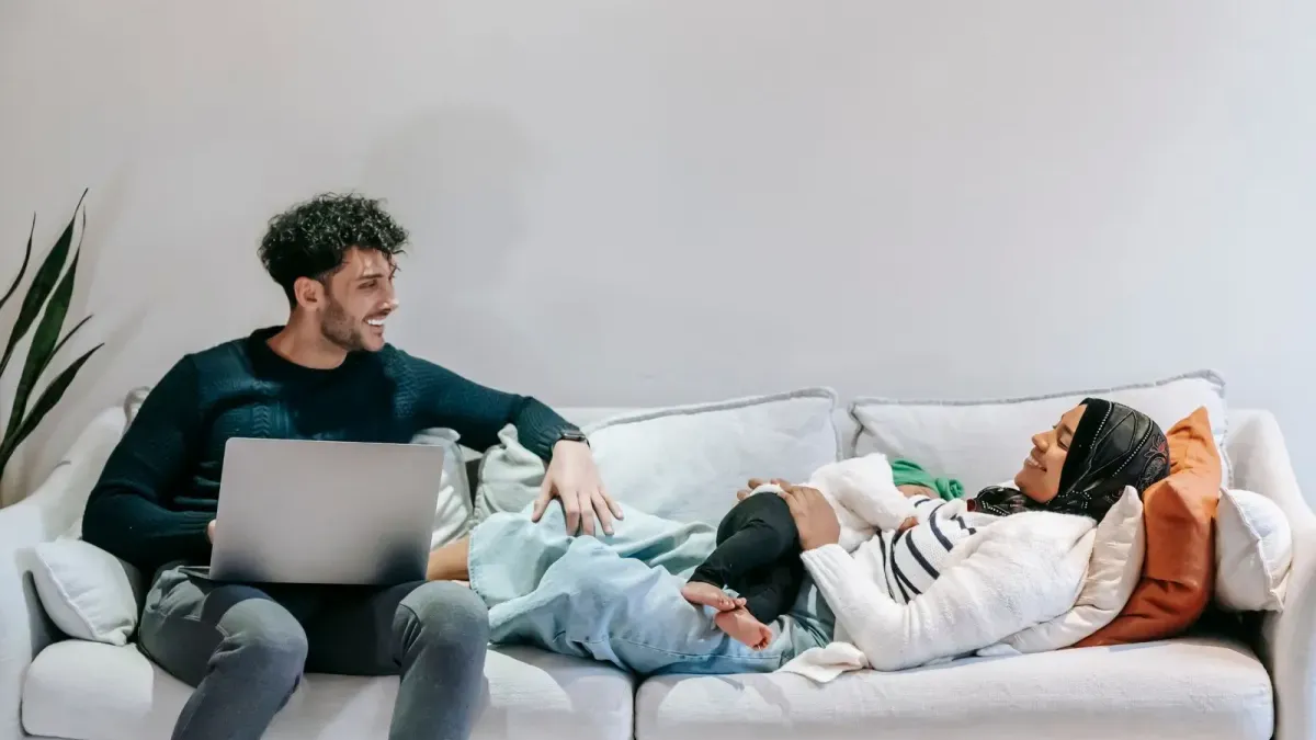 Paar mit Baby entspannt auf dem Sofa, einer arbeitet am Laptop