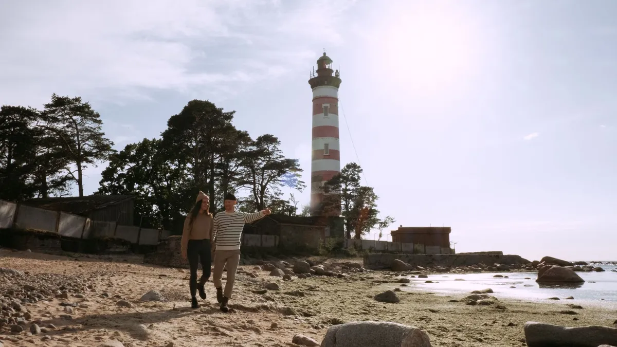 Paar spaziert an einem steinigen Strand, hinter ihnen ein rot-weißer Leuchtturm