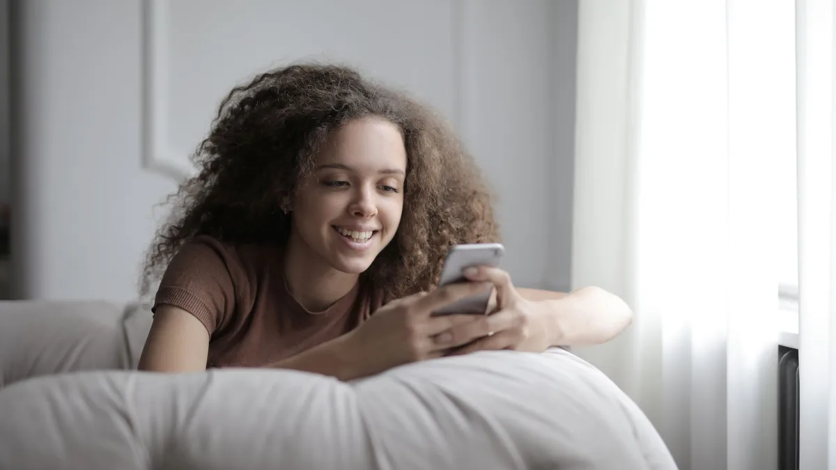 Lächelnde Frau liegt auf dem Sofa und schreibt auf dem Handy