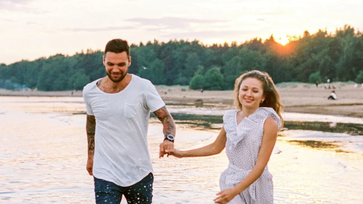 Lächelndes Paar läuft Hand in Hand durch flaches Wasser am Strand, im Hintergrund geht die Sonne unter