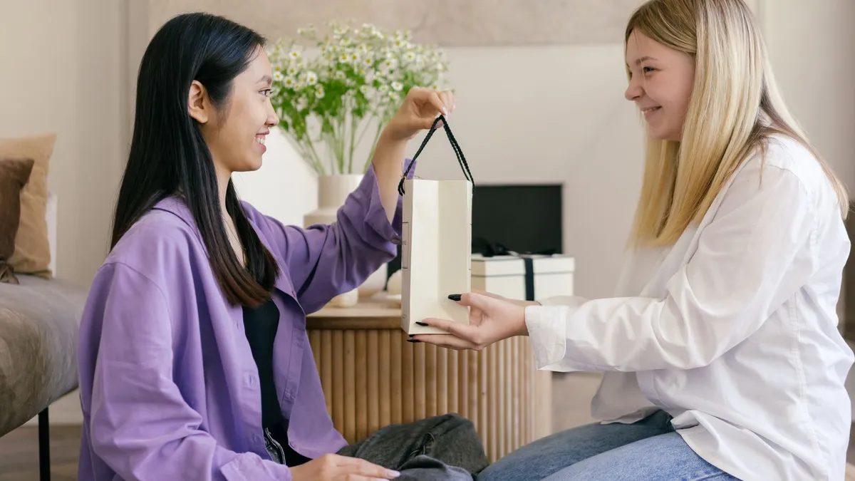 Zwei Freundinnen sitzen im Wohnzimmer, eine überreicht der anderen eine Geschenktüte