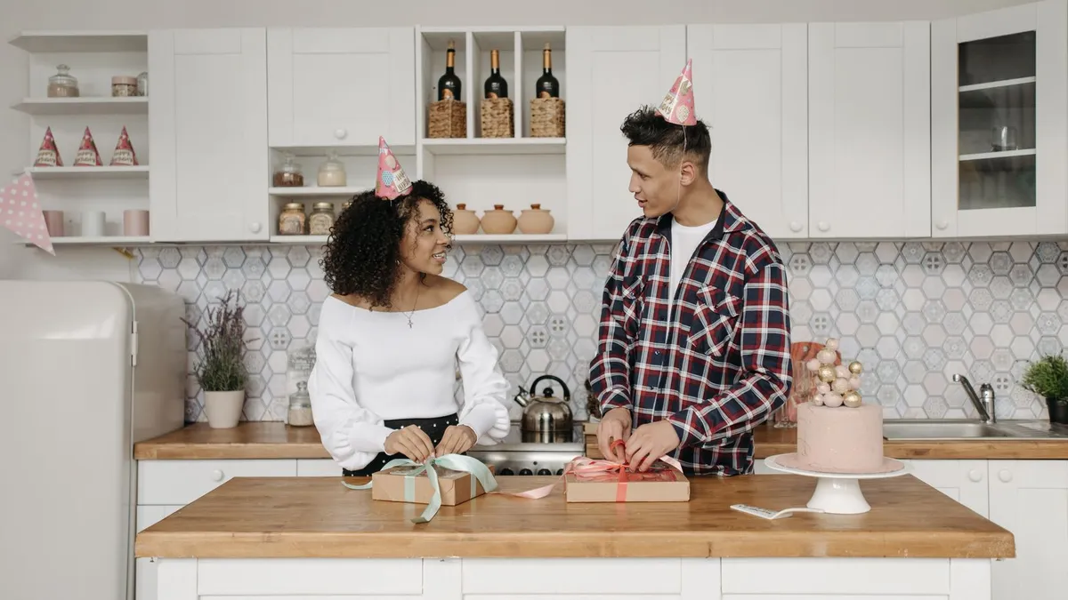 Paar mit Partyhüten packt in der Küche Geschenke ein, daneben steht eine Torte