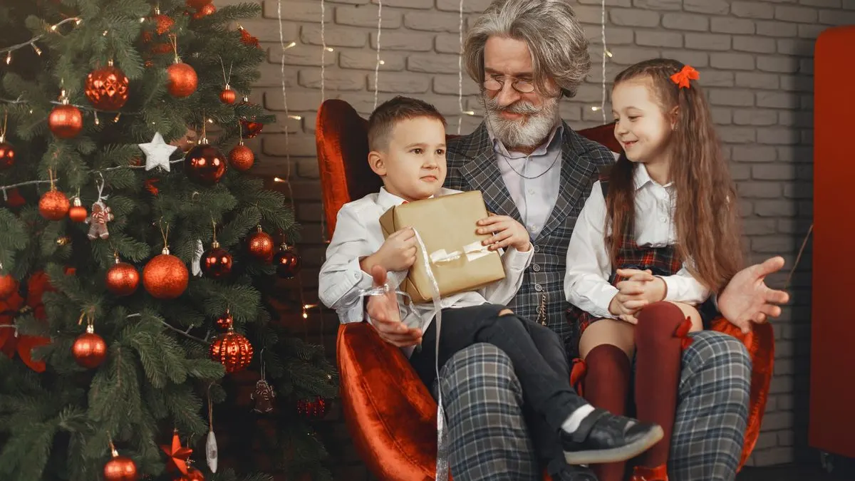 Großvater mit grauem Bart sitzt im roten Sessel vor dem Weihnachtsbaum, neben ihm zwei Enkelkinder, der Junge hält ein Geschenk