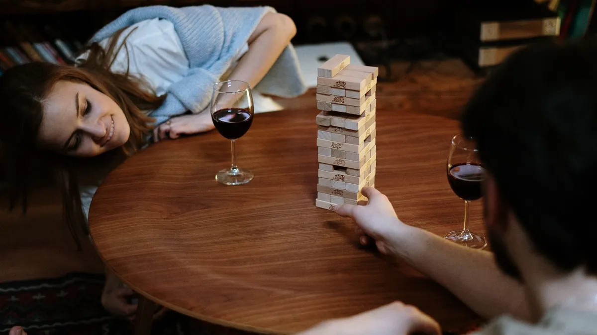 Paar spielt am Holztisch Jenga, daneben stehen zwei Gläser Rotwein