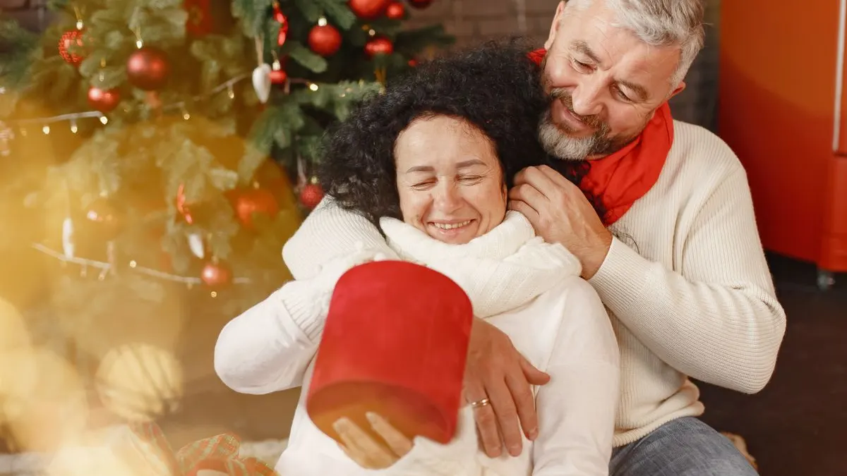 Älterer Mann umarmt lachend seine Partnerin, die vor dem Weihnachtsbaum eine rote Geschenkschachtel hält