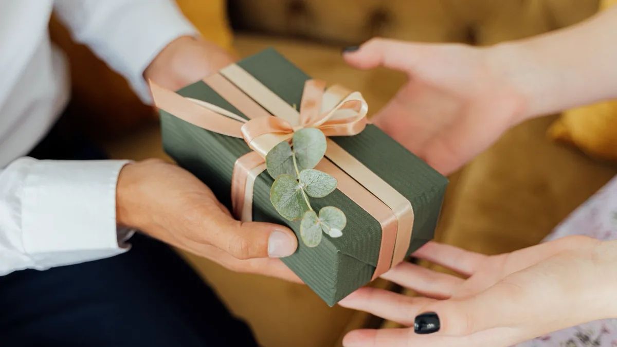 Hände überreichen ein grün verpacktes Geschenk mit Schleife und Eukalyptuszweig