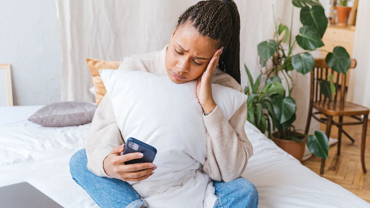 Junge Frau sitzt mit einem Kissen im Arm auf dem Bett und schaut bedrückt auf ihr Smartphone